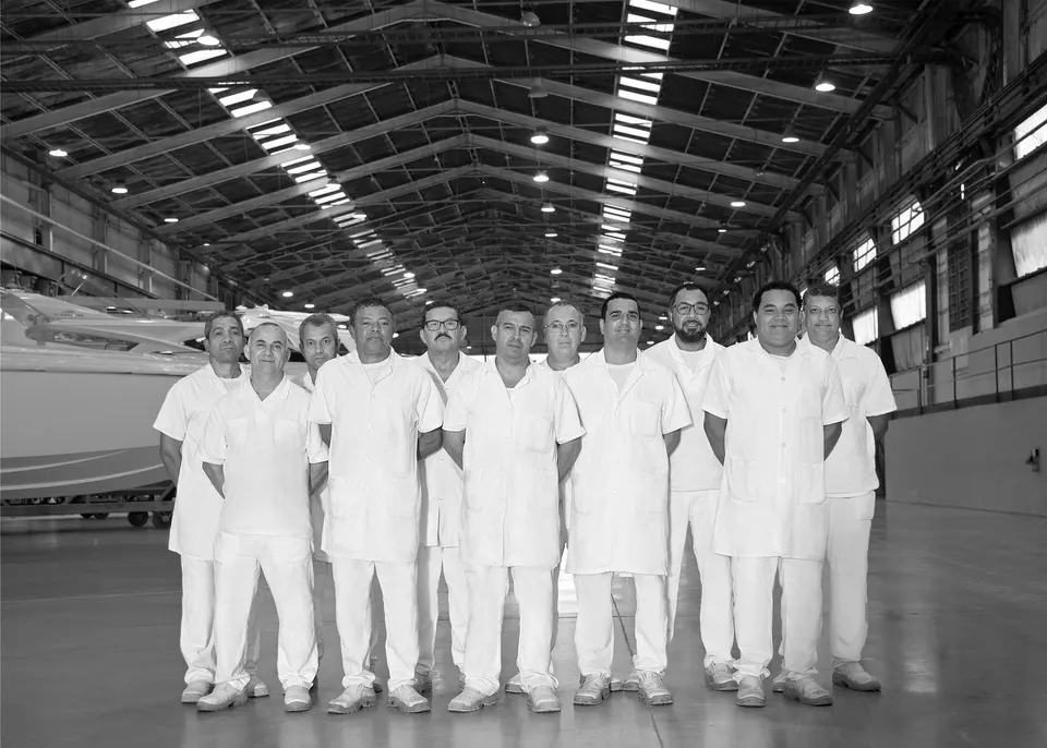 Dez artesãos do estaleiro, de uniforme branco, lado a lado em frente ao vão do galpão — em preto e branco